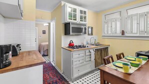 A compact kitchen with a microwave, kettle, and stove, a dining area with a table and chairs, and a small window with shutters.