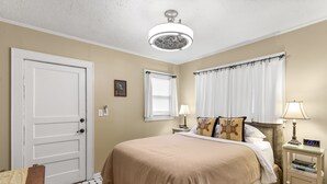 A hotel room with a bed, a nightstand, a lamp, a ceiling fan, a door, and a window with curtains.