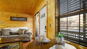 A cozy wooden cabin interior with a sofa, coffee table, and dining table set up. A sign on the wall reads "Gather together love and laughter forever."