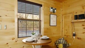 A wooden cabin interior with a round dining table set with plates and glasses, a window with blinds, a framed quote on the wall, and a shelf with speakers.