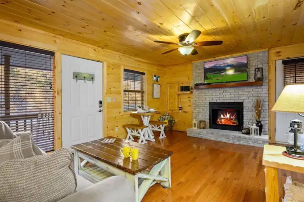 A cozy cabin interior with a wooden ceiling, a fireplace, a dining table, and a sofa.