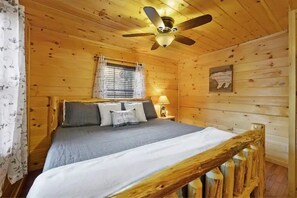 Um quarto de cabana de madeira com uma cama, um ventilador de teto, uma janela com cortinas e um abajur fixado na parede.