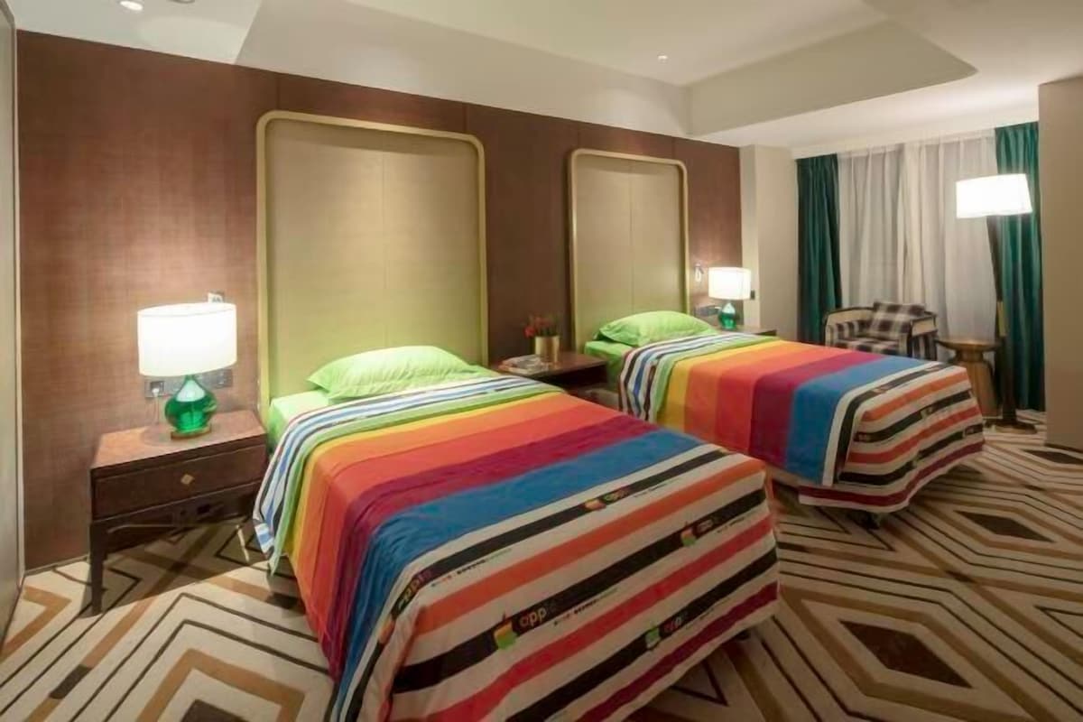 A hotel room with two beds, each with a striped bedspread, a nightstand with a lamp, a chair, and a large mirror on the wall.