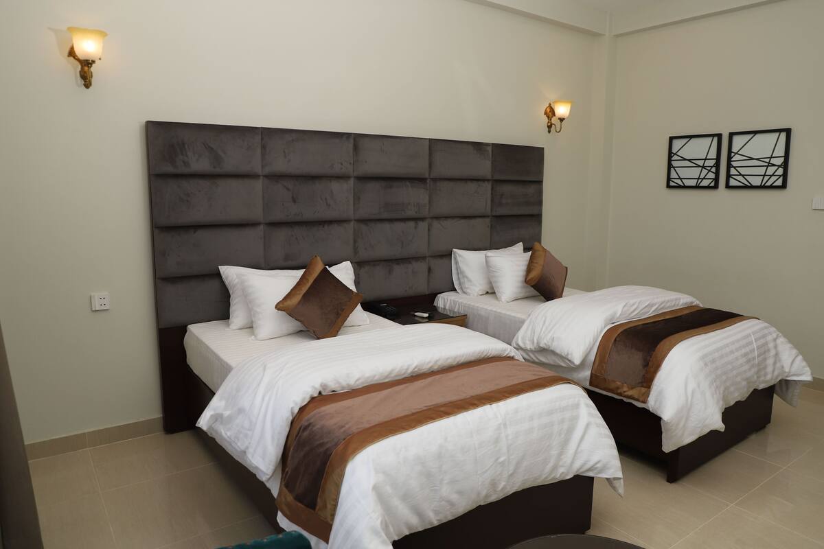 Une chambre d’hôtel avec deux lits, une grande tête de lit rembourrée, des lampes fixées au mur et deux œuvres d’art abstraites accrochées au mur.
