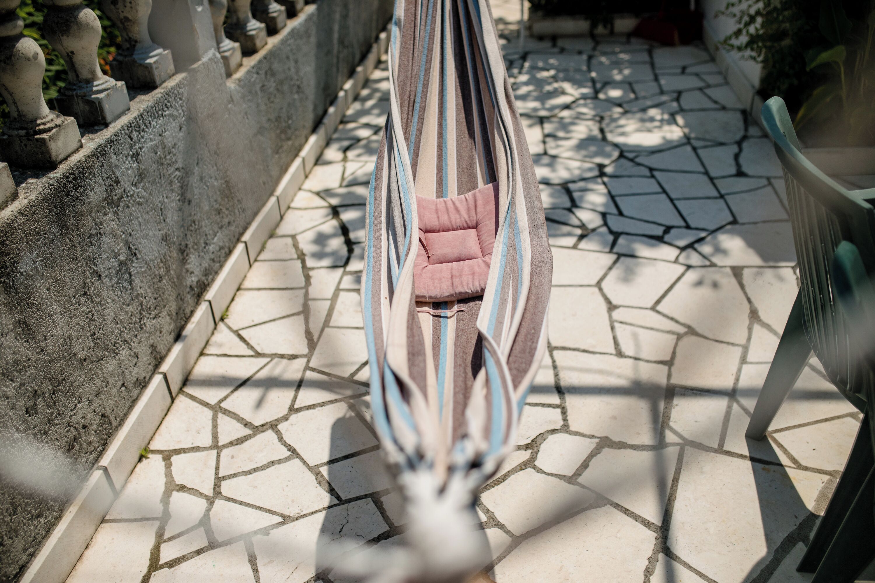 Een hangstoel met een roze kussen, geplaatst op een betegeld terras met een stenen muur en decoratieve balustrades.