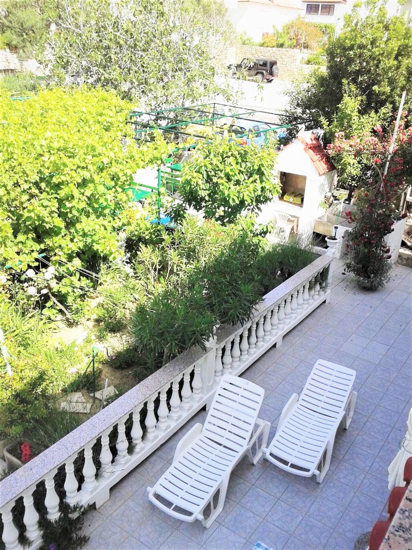 A balcony with white lounge chairs, a white balustrade, and a view of a garden with various plants and a building in the background.