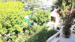 Ein Balkon mit weißen Lounge-Stühlen, einem weißen Geländer und Blick auf einen Garten mit verschiedenen Pflanzen sowie ein Gebäude im Hintergrund.