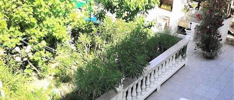 A balcony with white lounge chairs, a white balustrade, and a view of a garden with various plants and a building in the background.