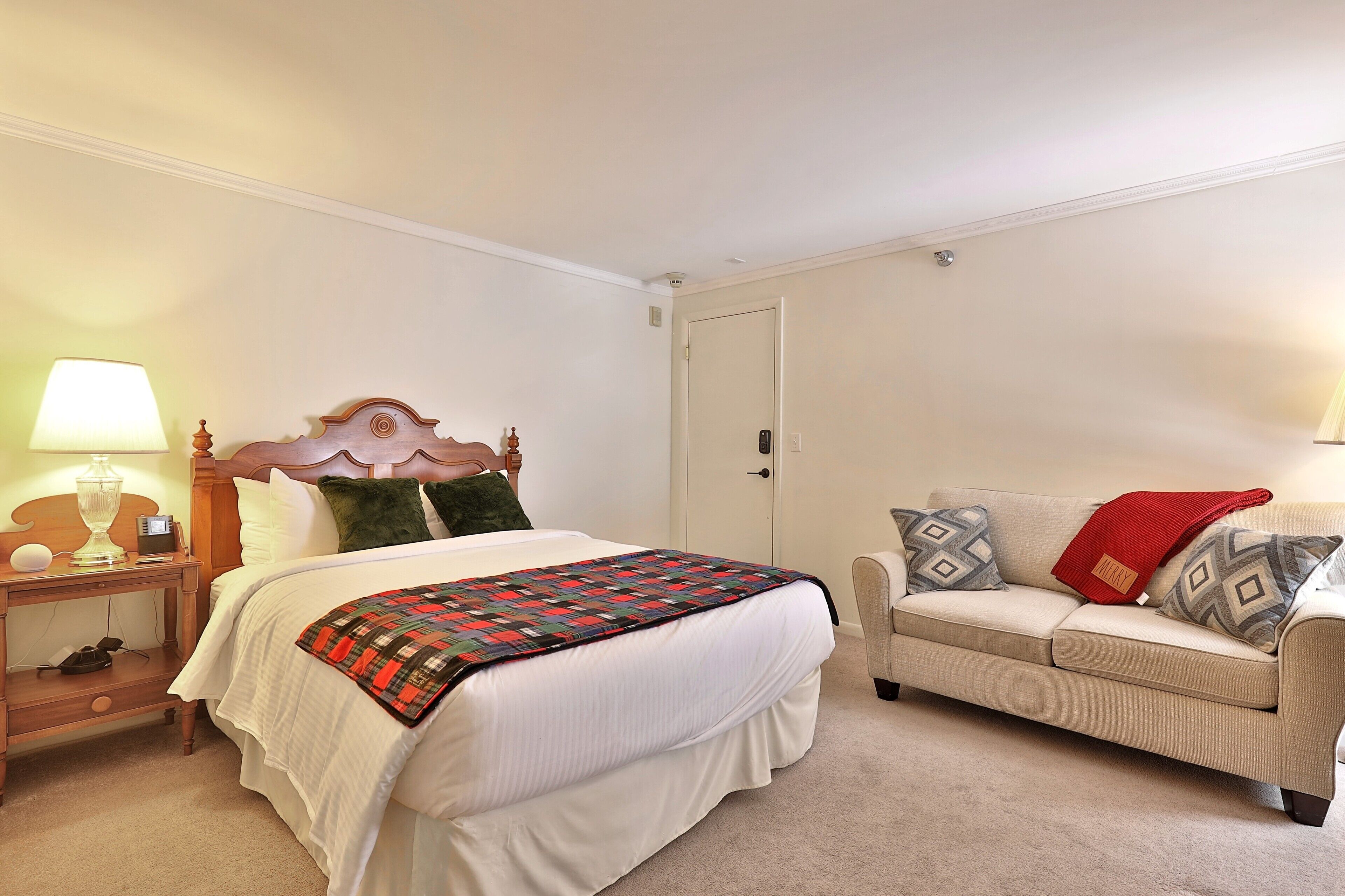 A bedroom with a bed, a nightstand, a bedside table, a lamp, a sofa, and a door.
