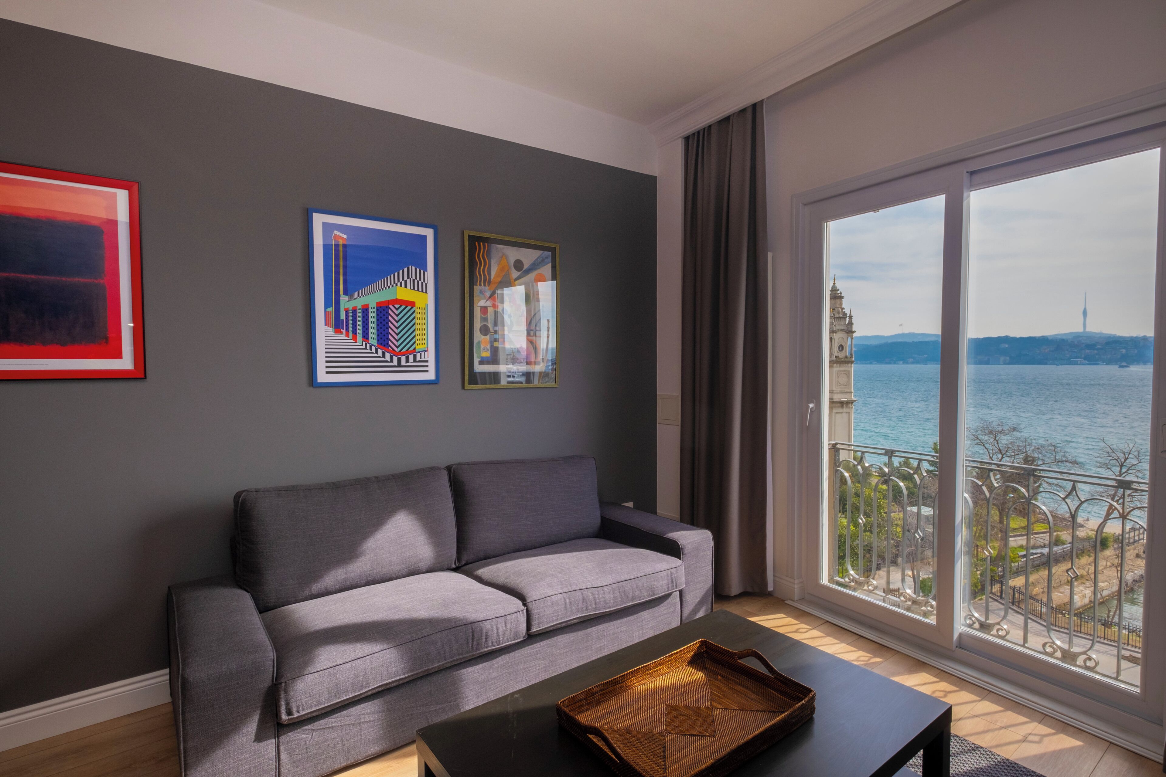 Una sala de estar moderna con un sofá gris, una mesa de centro negra y un ventanal con vistas a un paisaje urbano y una masa de agua.