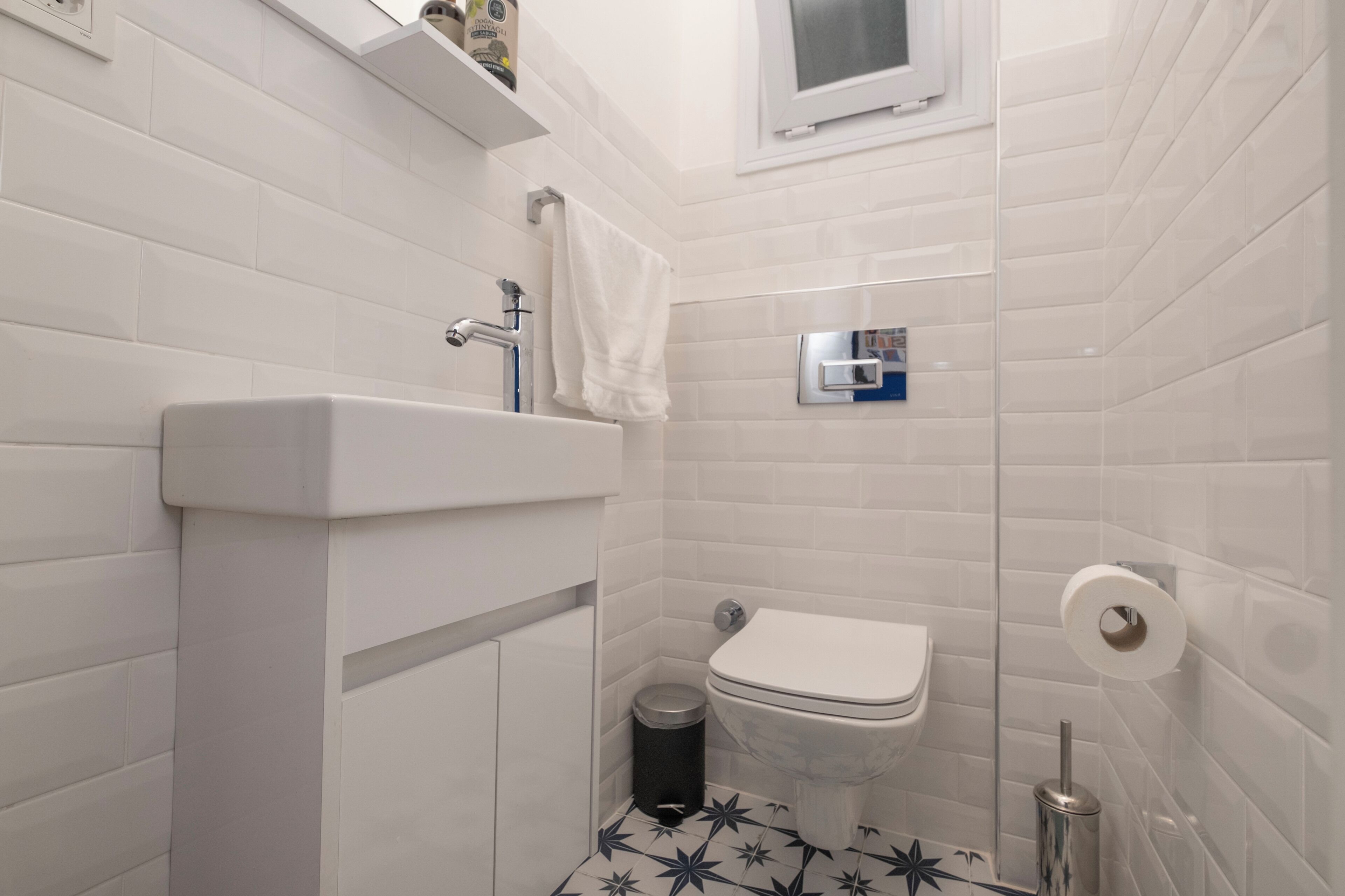 Une salle de bain avec des carreaux blancs, un lavabo blanc, des toilettes, une poubelle et une fenêtre située au-dessus des toilettes.