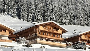 Eine Holzhütte mit Balkonen, umgeben von schneebedeckten Bäumen und wolkenlosem Himmel.