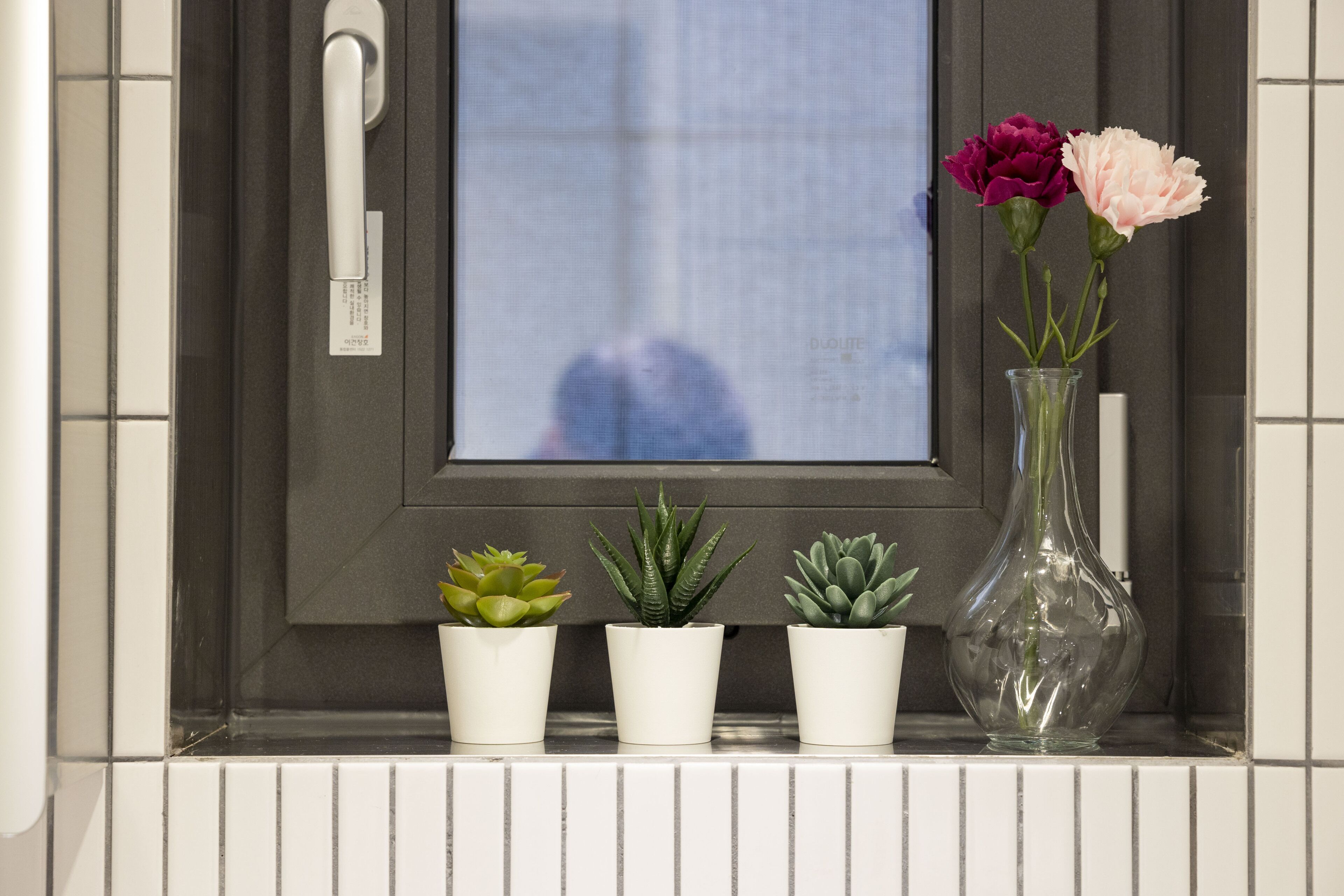 A window with a view of a building, three potted plants, and a vase with flowers.