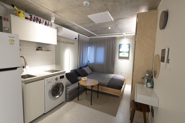 Un espacio habitable compacto con kitchenette, lavadora, cama y una pequeña mesa.