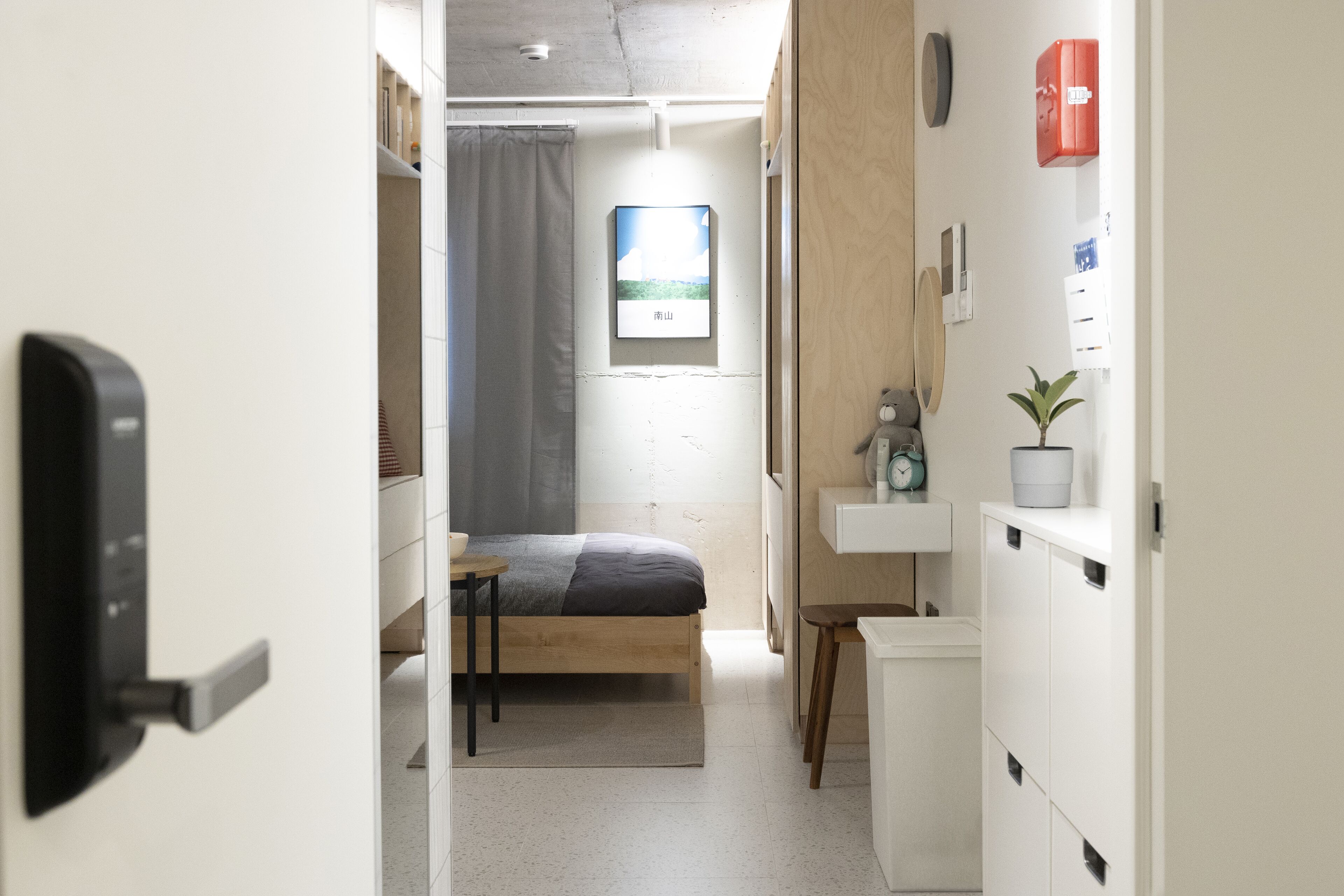 Un dormitorio pequeño con una cama, una mesita de noche y un escritorio. Se ve una ventana con vista a un edificio.