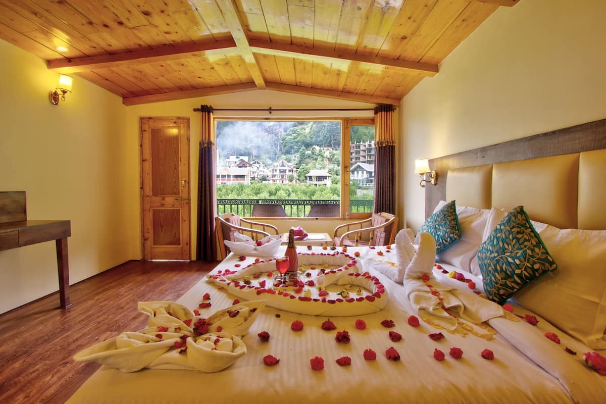 A hotel room with a wooden ceiling, a bed with rose petals, a balcony with chairs, and a view of buildings.