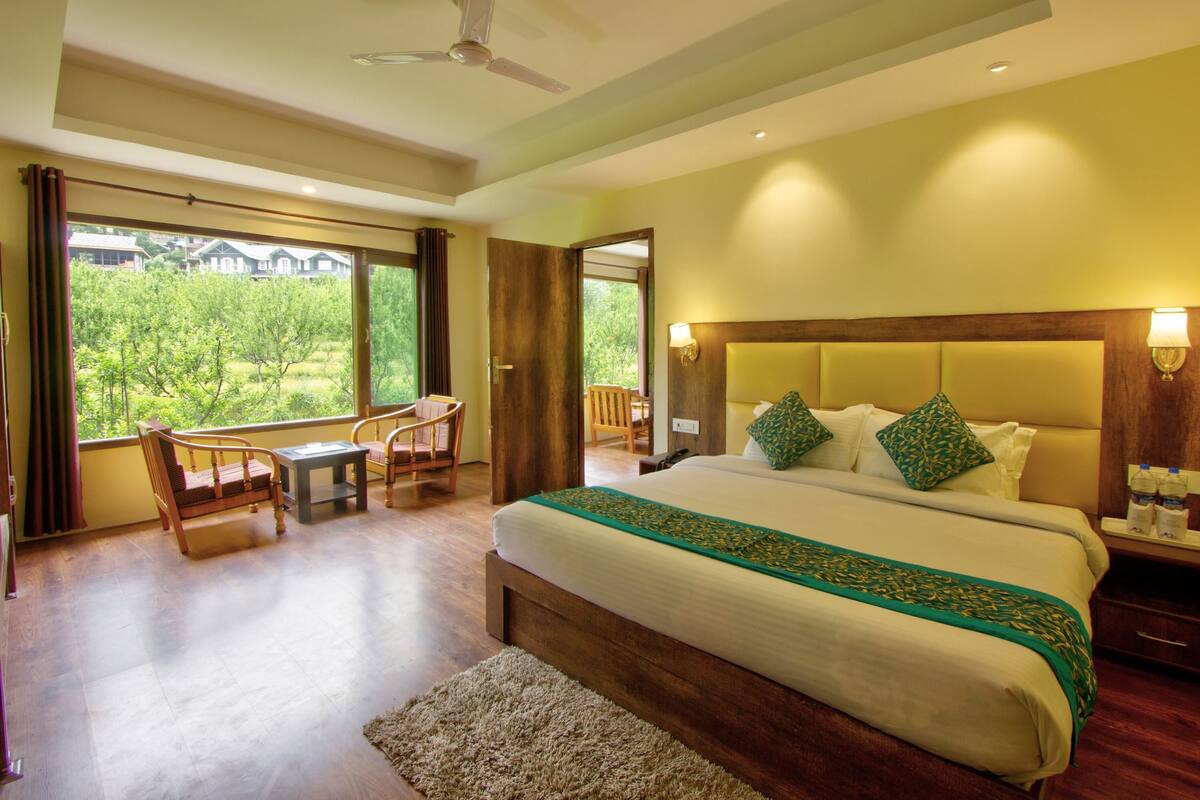 A bedroom with a large bed, a desk, two chairs, a window with a view, and a ceiling fan.