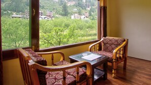A room with a large window offering a view of buildings and greenery. There is a wooden chair and a small table with a cushion. - Sunrise Villa Manali (Manali)