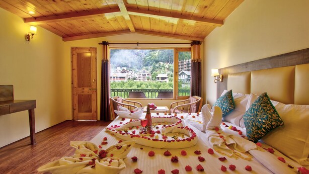 A hotel room with a wooden ceiling, a bed with rose petals and a bottle, a balcony with chairs, and a view of buildings. - Sunrise Villa Manali (Manali)