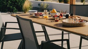 An outdoor dining area with a table set for breakfast, surrounded by chairs, and overlooking a scenic landscape. - Stylish Beach House 4 BR w/pool - 1 min from the beach! (Rhodes)