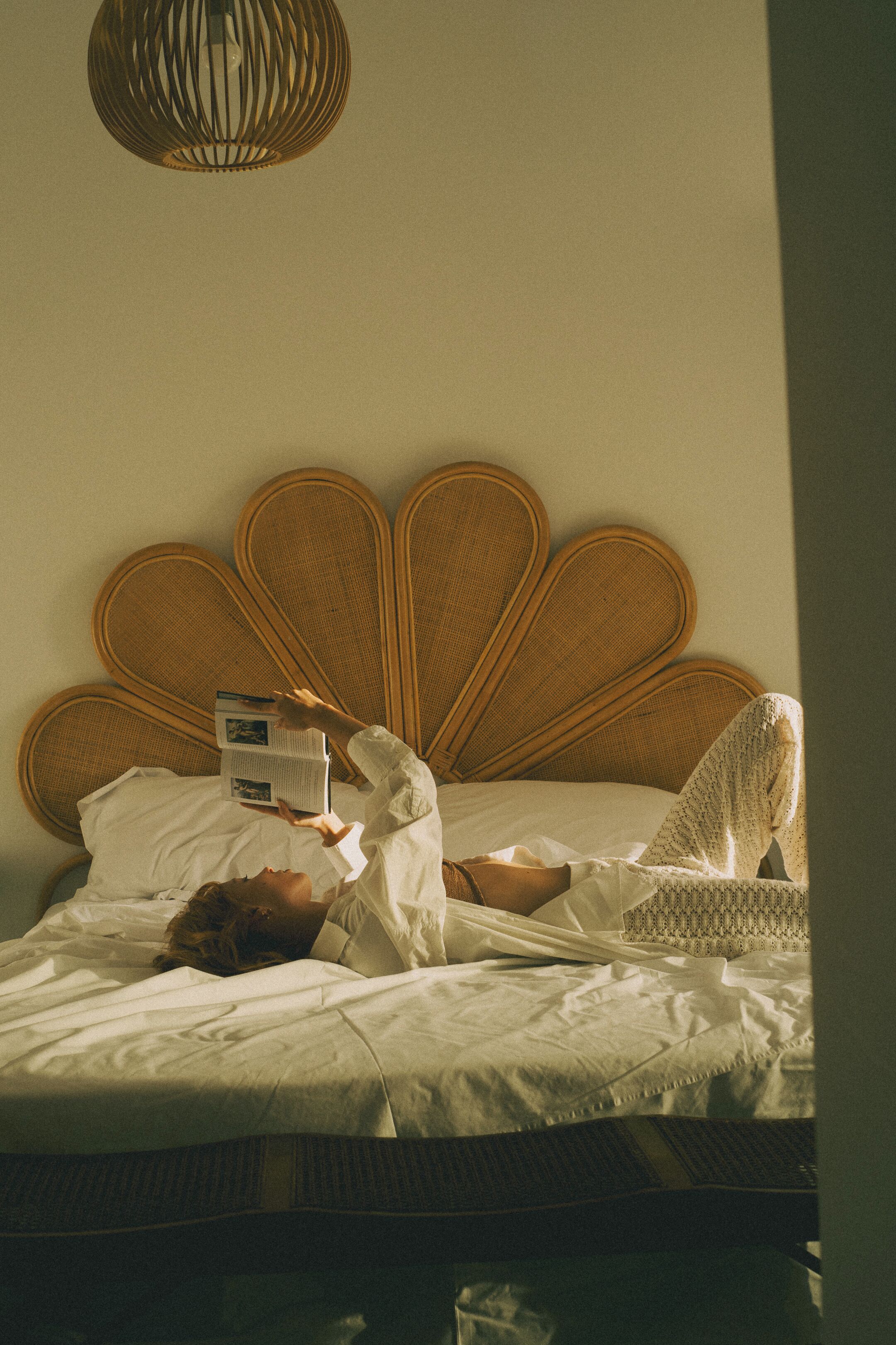 Een bed met een geweven hoofdbord, een persoon die erin ligt te lezen en een hangende lamp erboven.