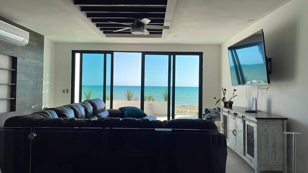Un salon moderne avec un grand canapé, un téléviseur fixé au mur et une vue sur la plage grâce à de grandes fenêtres.