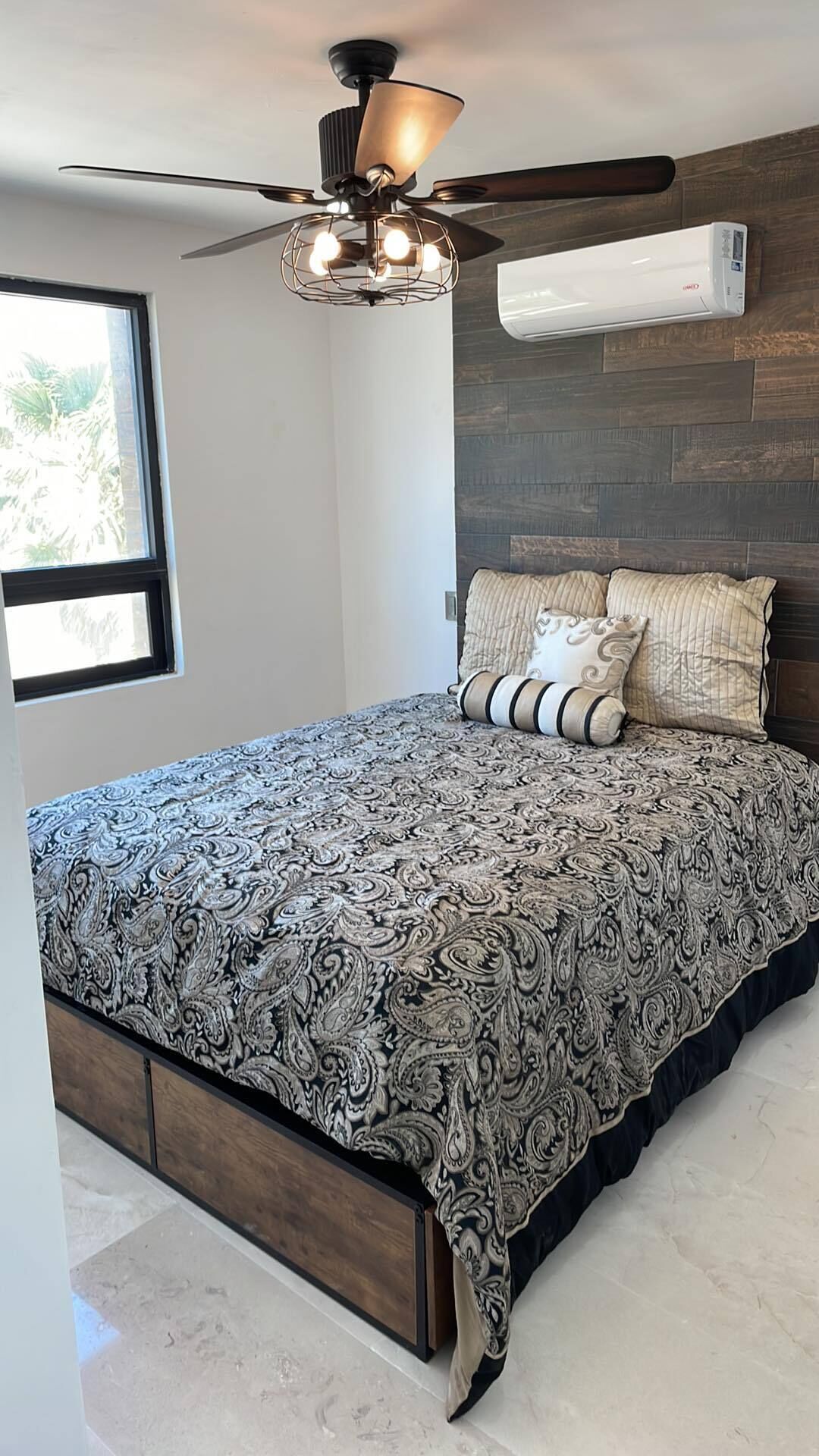 A bedroom with a large bed, a ceiling fan, an air conditioning unit, and a window with a view of greenery.