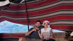 Un couple est assis sur un canapé, recouvert d’une couverture rouge et noire à motifs. On aperçoit une table avec une banane dessus, ainsi qu’une fenêtre donnant sur un paysage verdoyant.