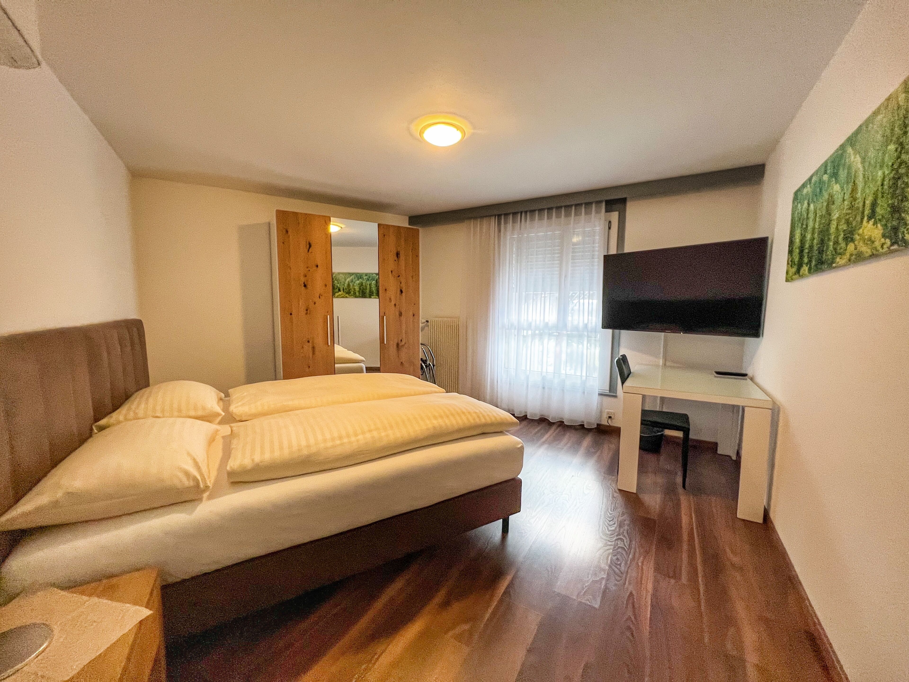 Ein Schlafzimmer mit Bett, Schreibtisch, Fernseher und einem Fenster mit Vorhängen.