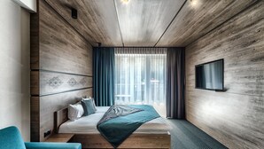 A modern hotel room with a wooden ceiling, a bed with a green bedspread, a blue armchair, and a TV mounted on the wall.
