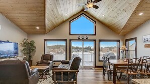 Amplia sala de estar con techo alto, vigas de madera, un televisor grande, sofás de cuero, una mesa de comedor con sillas y ventanas grandes que ofrecen vistas al exterior.