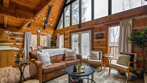 A cozy cabin interior with a wooden ceiling, a brown leather sofa, a round coffee table, and a large window with curtains.