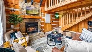 A cozy log cabin interior with a fireplace, wooden staircase, and comfortable seating areas.