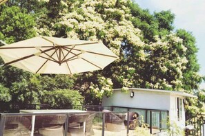 Un área de estar al aire libre con una mesa, sillas, un paraguas grande y un edificio con fachada de vidrio.