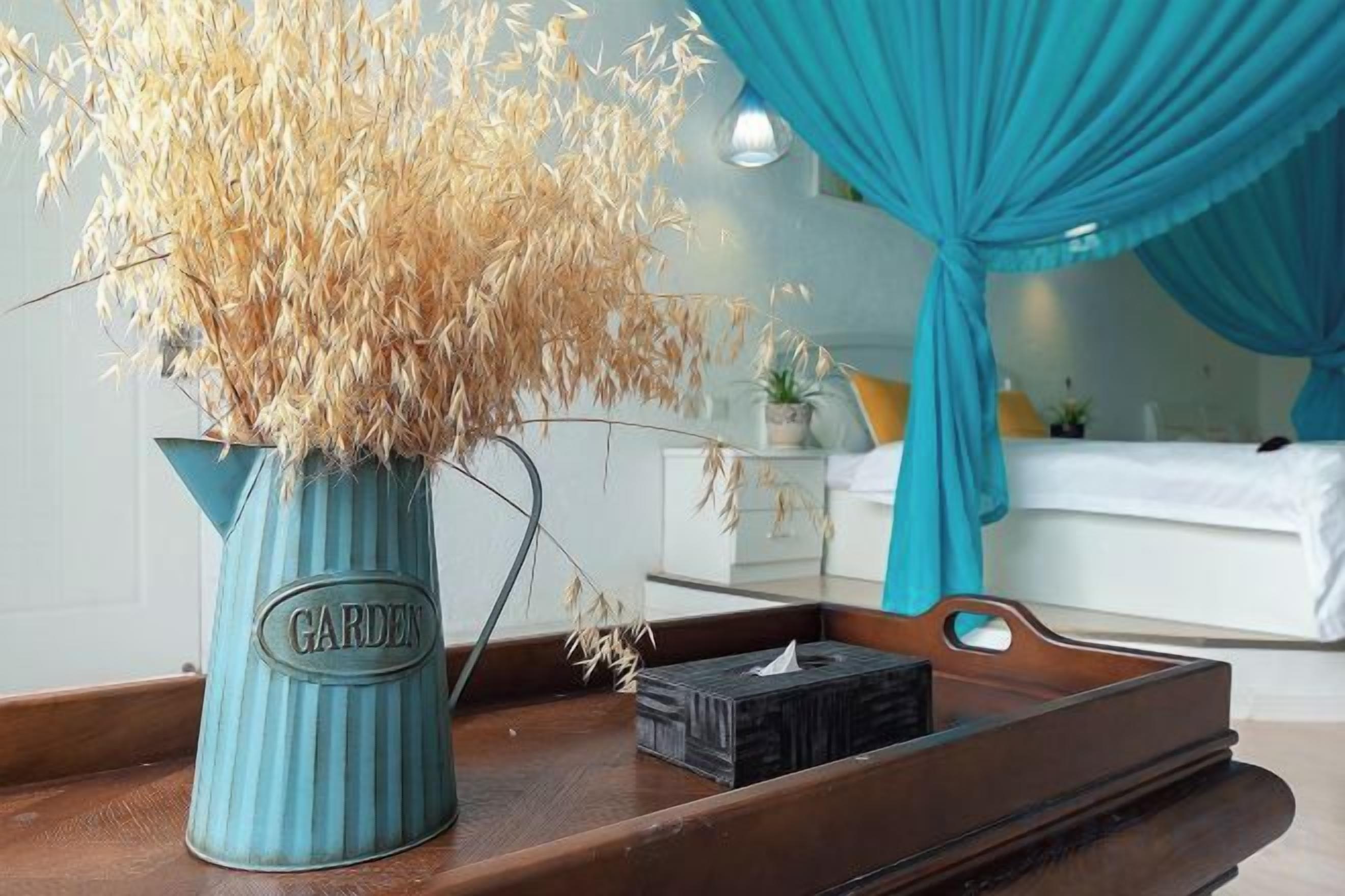 Un florero con plantas secas sobre una bandeja de madera en una habitación con cama y cortinas.