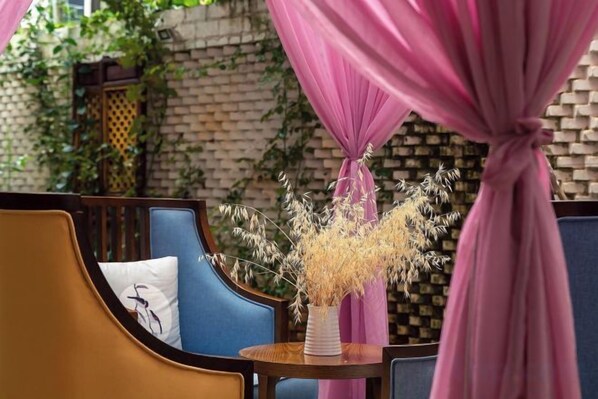 Un rincón de descanso con un banco acolchado, una mesa pequeña con un jarrón de flores secas y una pared de ladrillo con vegetación al fondo.