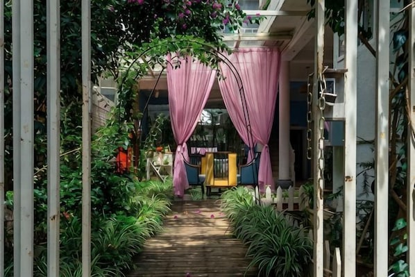 Un chemin menant à l’entrée d’un bâtiment, avec une terrasse en bois, entouré de verdure et d’une clôture blanche en piquets.