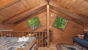 Un dormitorio abuhardillado de madera con una cama, un sofá y dos ventanas con vistas al verdor.