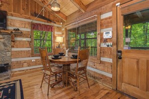 A wooden cabin interior with a dining table, chairs, a stone fireplace, a window with blinds, and a door with a keypad lock. - Perfect Cabin in the Woods! HOT TUB &  Game Room! (Sevierville)