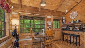 Interior de una cabaña de madera con una mesa de comedor, sillas, zona de bar y un ventanal con persianas.