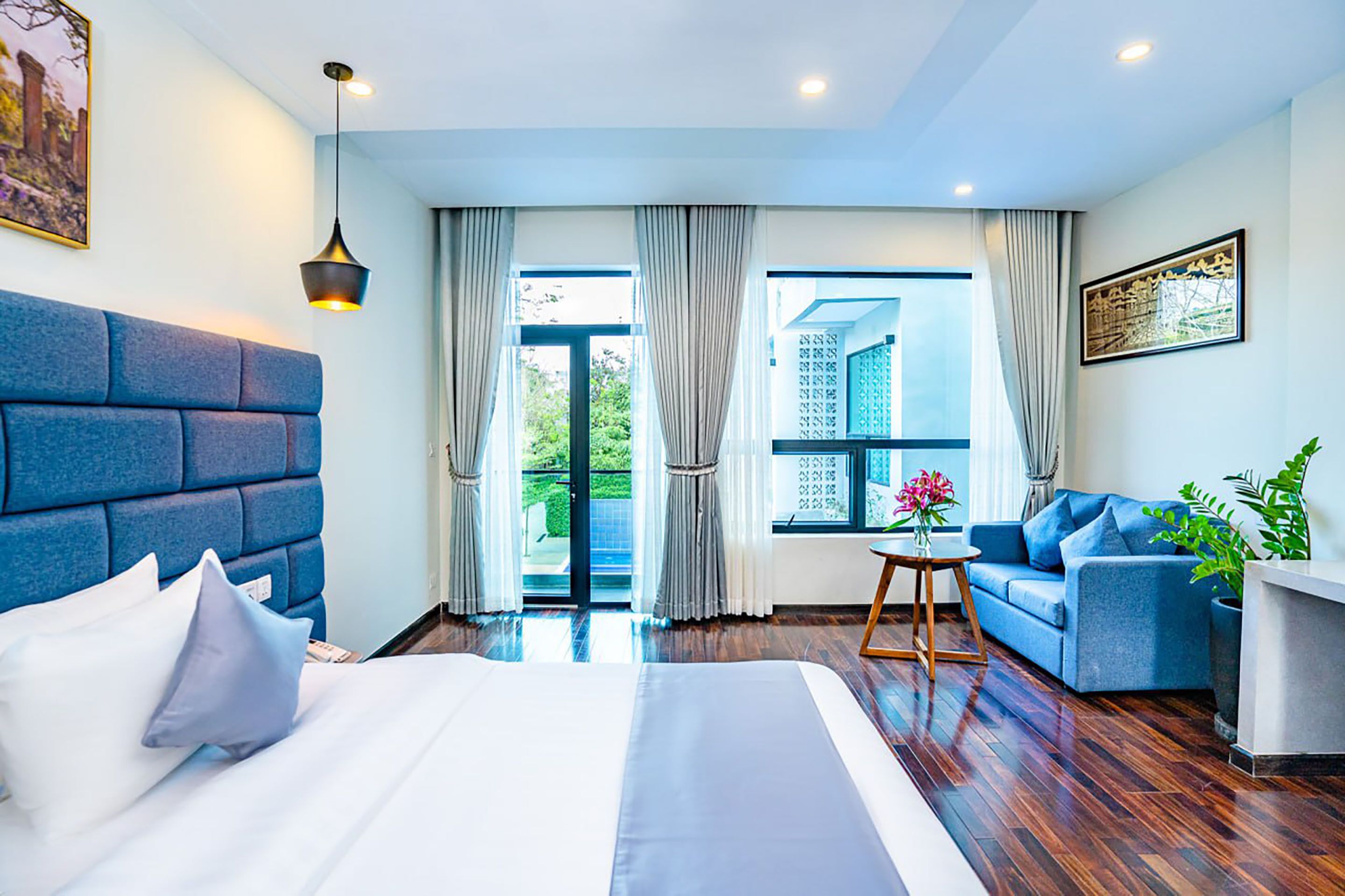 A modern hotel room with a large bed, a blue sofa, a small table with flowers, and a view of the outdoors through large windows.