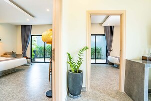 A modern hotel room with a large bed, a seating area with a sofa and armchairs, a dining table, and a potted plant.