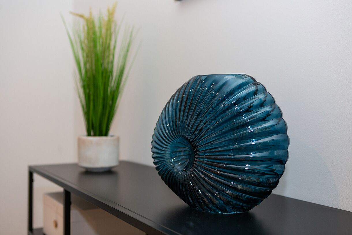 Un florero de vidrio azul verdoso con textura nerviosa sobre una mesa negra, junto a una maceta blanca con plantas verdes.