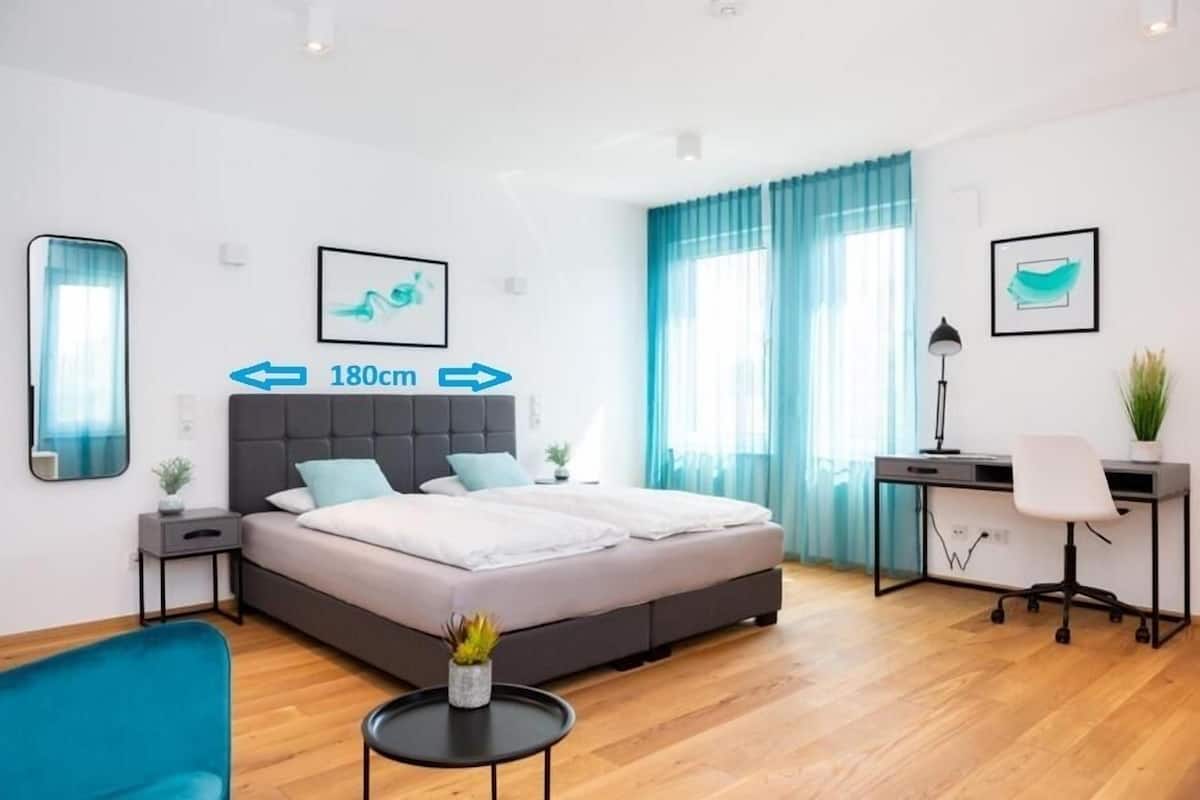 Una habitación de hotel moderna con una cama, un escritorio y una silla. Hay una mesita con una planta. La habitación tiene ventanas amplias con cortinas.