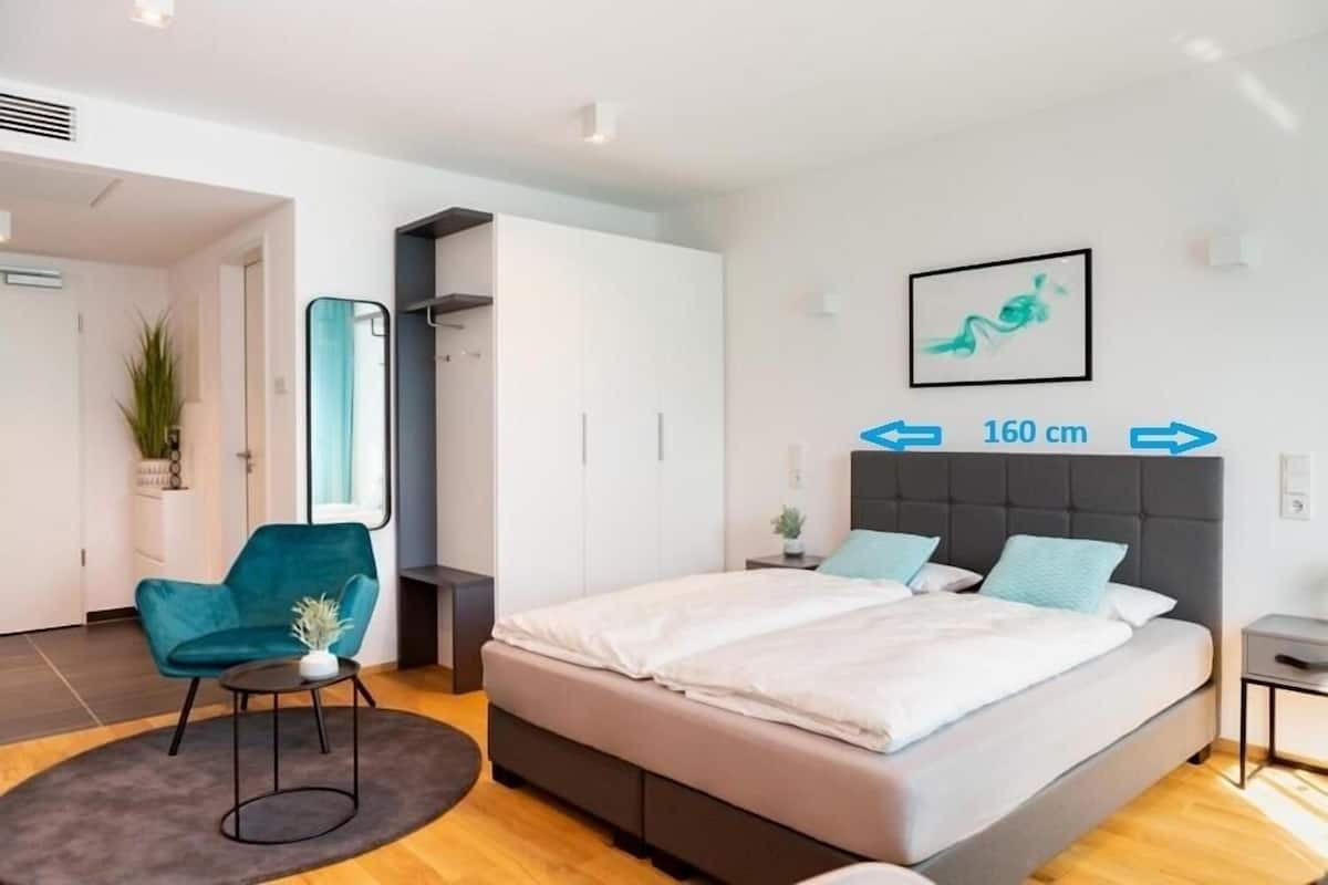 Un dormitorio moderno con cama, una silla y un espejo. La pared es blanca y tiene un cuadro enmarcado.