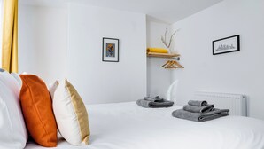 A hotel room with a neatly made bed, a small shelf with towels, and two framed pictures on the wall.