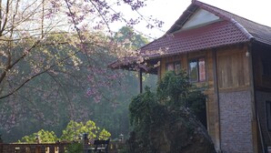 A traditional wooden house with a tiled roof, surrounded by lush greenery and flowering trees. - Farmstay Ban Bua (Moc Son)