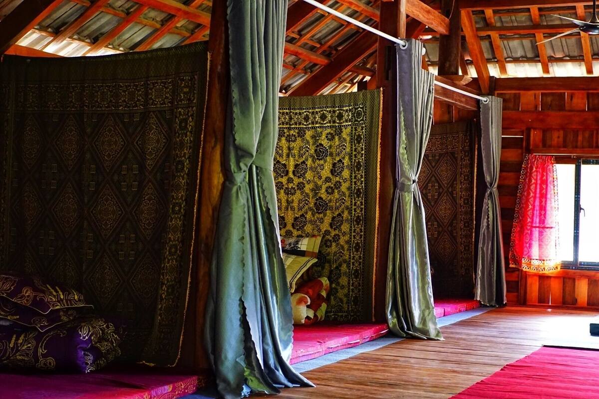 A room with a wooden ceiling and walls, a bed with a red and purple bedspread, and a window with a red curtain.