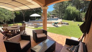 Uma área de pátio ao ar livre com mobiliário de vime, uma mesa com tampo de vidro e vista para uma piscina e jardim.