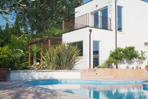 Un edificio moderno de dos pisos con piscina, rodeado de abundante vegetación y una pérgola de madera.